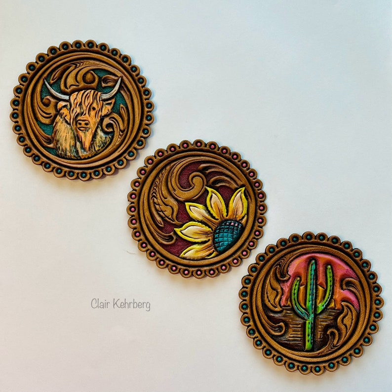 K&ouml;nnte beinhalten: Drei runde Lederuntersetzer mit aufwendigen Designs. Der erste Untersetzer zeigt eine braune und wei&szlig;e Highland-Kuh, der zweite eine gelbe Sonnenblume mit t&uuml;rkisfarbenen Akzenten und der dritte einen gr&uuml;nen Kaktus mit einem rosafarbenen Sonnenuntergang.
