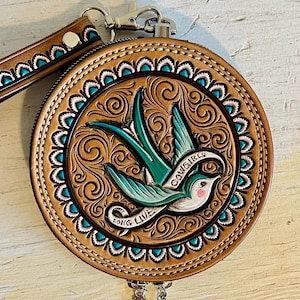 May include: Round, brown leather wallet with a turquoise and white border. The wallet features a detailed carving of a bird with the words "Cowgirls" and "Long Live". Includes a wrist strap and silver-tone hardware.