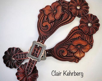 May include: A pair of brown leather spur straps with intricate floral designs and silver buckles. The straps are decorated with a repeating pattern of flowers and leaves. The buckles are silver and have a decorative design. The straps are attached to a pair of spurs, which are not visible in the image. Clair Kehrberg is printed on the bottom of the image.