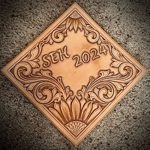 Può includere: Un pezzo quadrato di pelle con un intricato disegno floreale intagliato. Il centro del disegno presenta il testo "SEH 2024".