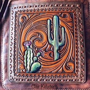 May include: A brown leather purse with a tooled design featuring a cactus and floral patterns. The design is framed by a stitched border. The purse has a signature "Clair Kehrberg" at the bottom.