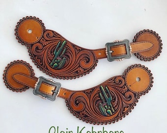 May include: A pair of brown leather cowboy spur straps with intricate floral designs and green cactus accents. The straps have silver buckles and are decorated with small beads.