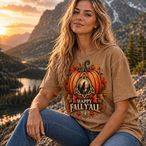 May include: A tan short-sleeve t-shirt featuring a graphic of a large orange pumpkin surrounded by autumn leaves and acorns. The design includes a central triangular emblem with a howling wolf silhouette and the text HAPPY FALL Y'ALL.
