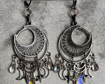 May include: Silver-toned chandelier earrings featuring crescent moon and star charms. The earrings have a filigree design with dangling teardrop and moon-shaped accents. One moon charm is yellow, the other is blue.