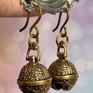 May include: A pair of antique gold-colored earrings. Each earring features a bell-shaped pendant with intricate embossed patterns. The earrings have a hook closure and are suspended from a decorative element.