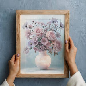 以下が含まれることがあります： ピンクのバラやその他の花が白い花瓶に入ったブーケが描かれた、額入りの花のプリント。 アートワークは水色の背景に配置され、天然木で額装されています。