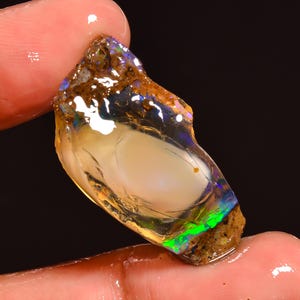 Könnte beinhalten: Ein roher Opal-Edelstein mit einem milchig-weißen Kern und schillernden Blitzen in Grün, Blau und Lila. Die raue Außenseite ist eine Mischung aus Braun und Beige. Der Edelstein wird zwischen zwei Fingern gehalten.