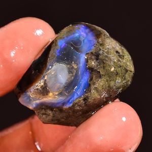 Könnte beinhalten: Ein roher Opal-Edelstein mit einer Mischung aus Farben. Der Stein hat eine dunkle, erdige Außenseite mit einem lebendigen blauen und weißen Inneren. Der Opal wird zwischen zwei Fingern gehalten.