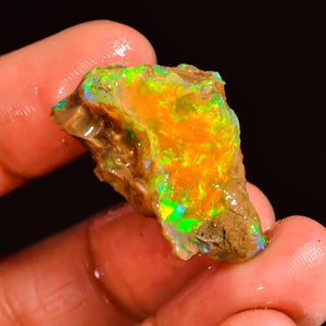May include: A vibrant, rough-cut opal gemstone. The stone displays a spectrum of colors, including bright orange, green, and blue, with a textured surface. The opal is held in a hand against a dark background.