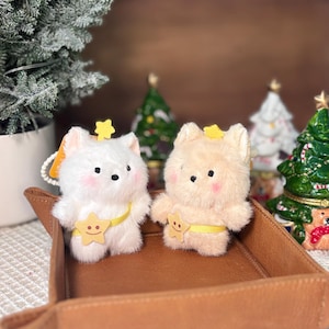 May include: Two plush bear-like figures, one white and one tan, each with a yellow star crown and a yellow star belly band. They sit in a brown leather tray. Christmas trees are in the background.