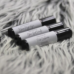 May include: Three perfume oil roll-on bottles with black caps and white labels, resting on a fluffy, gray surface. The labels read "Bloom Hypnotique", "Clean Girl Vanilla", and "Pink Cashmere". The bottles are from "Daisy & Blush".