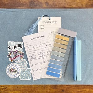 May include: A reading list bookmark with a light blue tassel, a book review card, a set of adhesive page markers in shades of blue and tan, a clear ruler, and two light blue pens, accompanied by four decorative stickers with book-themed text.