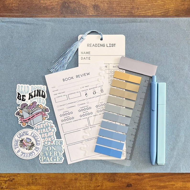 May include: A reading list bookmark with a light blue tassel, a book review card, a set of adhesive page markers in shades of blue and tan, a clear ruler, and two light blue pens, accompanied by four decorative stickers with book-themed text.