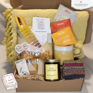 Pode incluir: Uma caixa de presente com tema amarelo, contendo uma caneca, uma vela, saquinhos de chá, meias e artigos de papelaria. A vela diz "Sun Shower". A caixa também inclui um adesivo com tema de livro e um pequeno livro que diz "A Novel Escape".