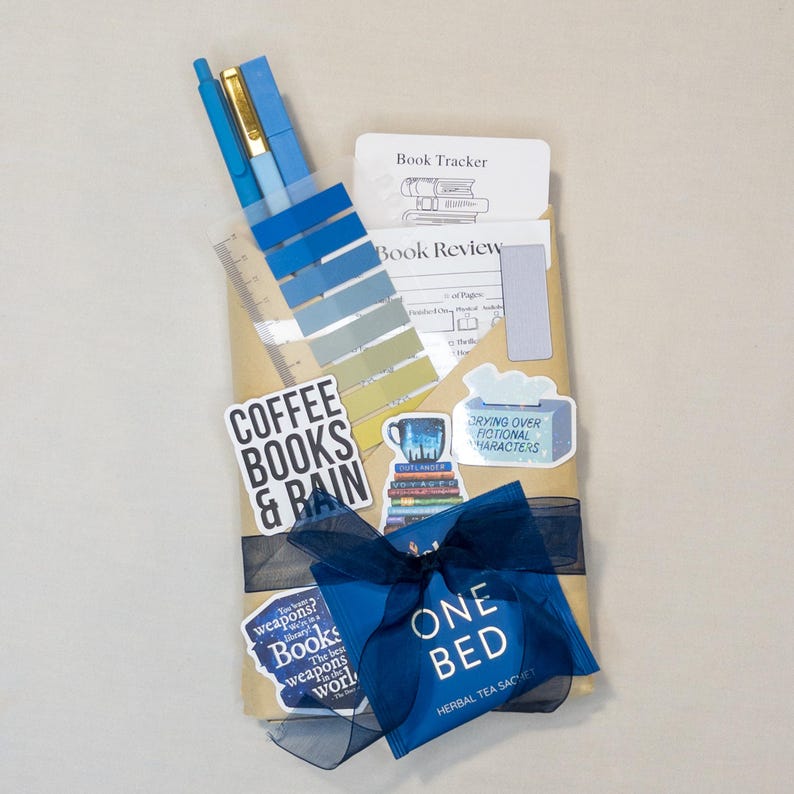 Puede incluir: Un juego de regalo envuelto en papel marr&oacute;n, atado con una cinta y un lazo azul marino. El juego incluye bol&iacute;grafos, una regla, pegatinas con texto como "Coffee Books & Rain" y tarjetas tem&aacute;ticas de libros.