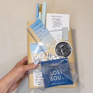 Puede incluir: Un paquete de regalo con una variedad de art&iacute;culos de papeler&iacute;a. Incluye bol&iacute;grafos azules, notas adhesivas, pegatinas y una bolsita de t&eacute;. El paquete est&aacute; decorado con pegatinas que dicen "Books are Magical" y "Lost Soul".