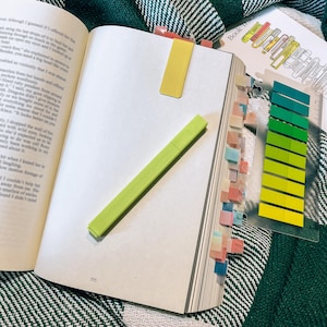 May include: An open book with a lime green highlighter resting on the page, a yellow magnetic bookmark attached to the top, and a clear plastic sheet featuring a vertical row of ten adhesive index tabs in shades of green and yellow.