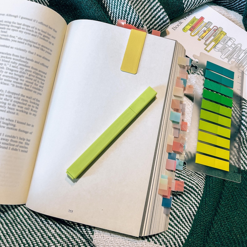 May include: An open book with a lime green highlighter resting on the page, a yellow magnetic bookmark attached to the top, and a clear plastic sheet featuring a vertical row of ten adhesive index tabs in shades of green and yellow.