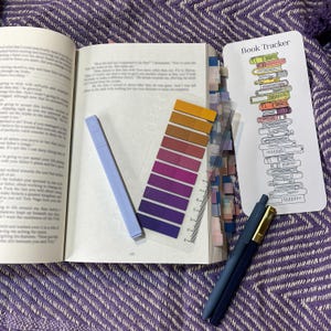 Puede incluir: Un libro abierto con marcadores de p&aacute;gina coloridos, un resaltador azul claro, una regla transparente con muestras de color, un marcador que dice "Book Tracker" y dos bol&iacute;grafos azules con detalles dorados, todo sobre una superficie morada estampada.