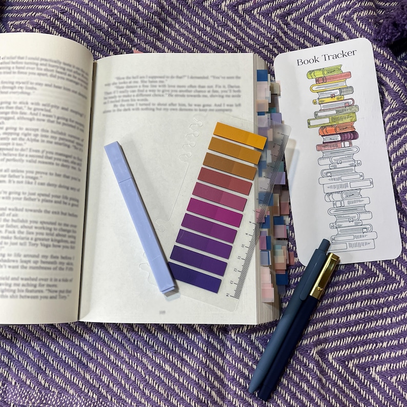 Puede incluir: Un libro abierto con marcadores de p&aacute;gina coloridos, un resaltador azul claro, una regla transparente con muestras de color, un marcador que dice "Book Tracker" y dos bol&iacute;grafos azules con detalles dorados, todo sobre una superficie morada estampada.