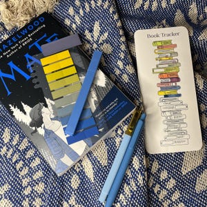 May include: A book titled Mate by Hazelwood, a set of adhesive page markers in yellow and blue gradients, two blue pens, and a white bookmark with a vertical illustration of a stack of books and the text Book Tracker printed at the top.