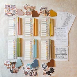 May include: A collection of stationery items, including pens, sticky notes, stickers, and bookmarks. The stickers feature phrases like "Hello Fall" and "Bookish Head." The pens come in various colours, and the bookmarks are heart-shaped.