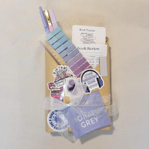 Puede incluir: Un juego de regalo para amantes de los libros, envuelto en papel marr&oacute;n y atado con una cinta blanca. Incluye bol&iacute;grafos, notas adhesivas, una regla, pegatinas y una bolsita de t&eacute;. Las pegatinas tienen el texto "Emotionally A Fictional", "Listening Audiobooks Reading" y "Moral Grey".