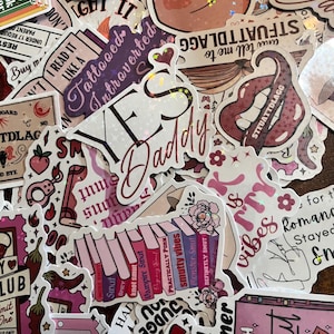 May include: A collection of assorted holographic stickers featuring various designs, including books with titles like 'Smutty Vibes', 'Monster Smut', and 'Definitely Smut', alongside text-based stickers reading 'Yes Daddy' and 'Tattooed Introverted'.