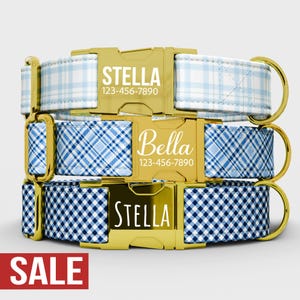 May include: Three personalized dog collars stacked on top of each other. The collars have gold-colored buckles and D-rings. The collars are light blue and white plaid, blue plaid, and blue and white gingham. Each collar has a name and phone number.