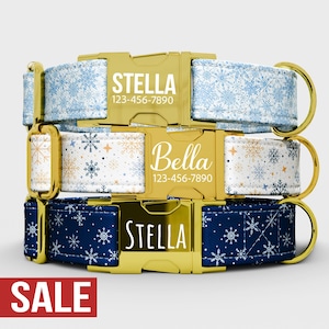 May include: Three dog collars with gold-colored buckles and D-rings. The collars feature snowflake patterns in blue, white, and navy. Each collar has a name and phone number printed on the buckle. The word "SALE" is in a red rectangle.