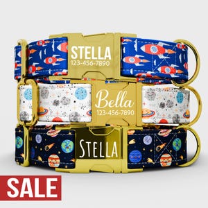 May include: Three dog collars with gold-colored buckles. The top collar is blue with red rockets. The middle collar is white with planets and smiling clouds. The bottom collar is navy with planets and stars. Each collar has a name and phone number.