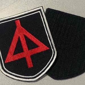 Może przedstawiać: Czarna naszywka z czerwonym trójkątnym wzorem i białym obramowaniem. Naszywka znajduje się obok oddzielnego kawałka czarnej tkaniny. Wzór to stylizowany kształt geometryczny.