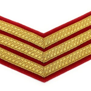 May include: A red chevron-shaped patch with three gold embroidered stripes. The stripes are detailed with a repeating pattern. The patch is likely a military or service rank insignia.