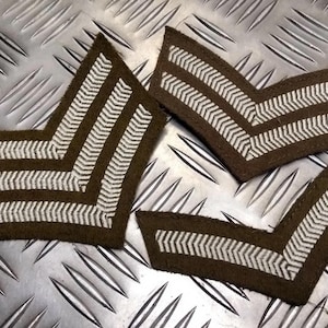 May include: Three olive green chevron patches with white zigzag stripes. The patches are arranged on a textured, metallic surface. These military-style patches are likely for uniforms or other apparel.