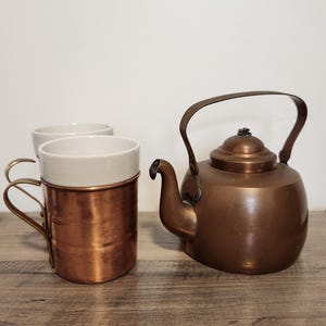 May include: A copper kettle and a copper mug with a white ceramic interior sit on a wooden surface. The kettle has a curved handle and a spout. The mug has a handle and a white interior. The items are vintage and have a rustic appearance.