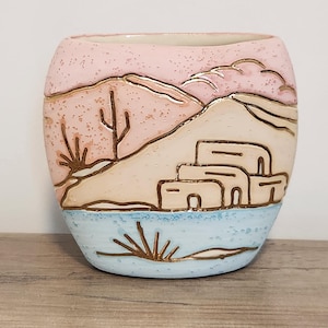 May include: A ceramic planter with a rounded square shape featuring a desert landscape relief. The design includes a pink sky, beige hills, a blue foreground, and gold-outlined cacti and adobe-style buildings with a speckled texture.