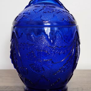 May include: A cobalt blue glass jar with a lid. The jar has a textured surface with a leaf and berry design. The jar is sitting on a wooden surface.