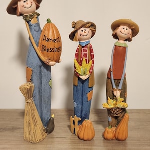 May include: Three whimsical scarecrow figurines. The first holds a pumpkin with "Harvest Blessings" text, the second holds corn, and the third holds a basket of corn. All wear hats, overalls, and have smiling faces. A broom and pumpkins are also present.