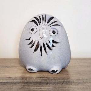 May include: A rounded, light blue ceramic owl figurine featuring hand-painted black details. The design includes two circular eyes, a central beak, and radiating black brushstrokes around the face, with two small black feet at the base.