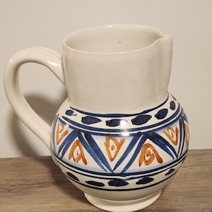 May include: A white ceramic pitcher with a rounded base and a curved handle. The pitcher features a blue and orange geometric pattern around the body. The top portion of the pitcher is plain white.