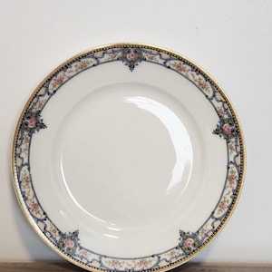 May include: A round white porcelain dinner plate featuring a decorative border with pink rose clusters, blue scrollwork, and a gold-trimmed edge. The center of the plate is plain white, and the rim is adorned with a repeating floral and geometric pattern.