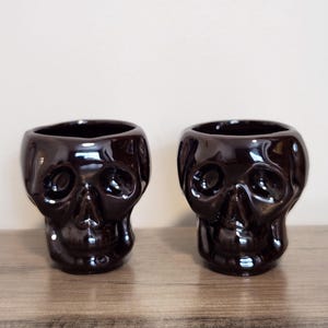 May include: Two identical dark brown ceramic mugs shaped like human skulls. Each mug features a glossy finish with molded eye sockets, a nasal cavity, and a defined jawline. The vessels have an open top and a smooth, reflective surface texture.