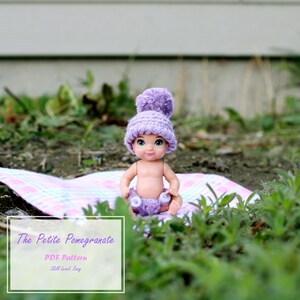 May include: A doll wearing a purple crocheted hat and diaper sits on a pink and white plaid blanket. The doll is in a natural outdoor setting with green grass and foliage. The text "The Petite Pomegranate" is in the lower left corner.