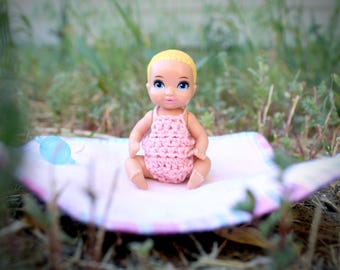 May include: A small baby doll with blonde hair and blue eyes, wearing a pink crocheted outfit. The doll is sitting on a pink and white patterned mat, with a blue bottle nearby. The scene is set outdoors with green grass in the background.