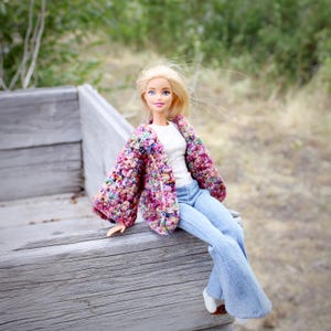 May include: A doll dressed in a multi-coloured crochet jacket, white top, and light blue flared jeans. The jacket has pink, purple, and other hues. The doll is seated on a weathered wooden surface.