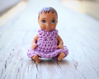 May include: A small doll with dark skin and brown hair wearing a handmade lavender crochet dress. The doll is sitting on a weathered wooden surface, showcasing the texture of the wood.