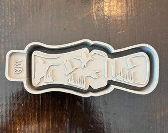 May include: A light gray cookie cutter in the shape of a duck call. The design includes a hand holding the call, with flying ducks and reeds. The cookie cutter is on a dark brown wooden surface.