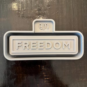 May include: A light gray 3D-printed object with the word "FREEDOM" in capital letters. The object has a rectangular shape with a smaller rectangular section on top, featuring abstract symbols. The object is on a dark brown surface.