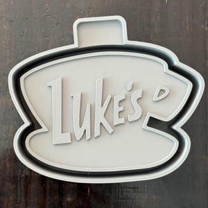 以下が含まれることがあります： 「Luke's」の文字が入った、コーヒーカップ型のライトグレーのクッキーカッター。 黒いアウトラインとハンドルが付いています。 背景は濃い木材の表面です。