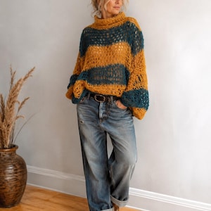Puede incluir: Un suéter holgado de manga larga con una textura de punto abierto y grueso, con rayas horizontales alternas en amarillo mostaza y azul cerceta oscuro. El suéter tiene un cuello alto doblado y se combina con jeans azules con dobladillo.